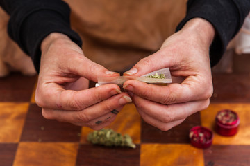 Closeup shot of hands pinching a cannabis filled rolling paper starting to roll it into a joint. Weed bud and grinder on the checkered table below.