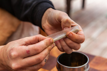 Selective focus closeup shot of tattooed hands twisting a paper with ground marijuana preparing a cannabis below a metal cylinder over a checkered table.