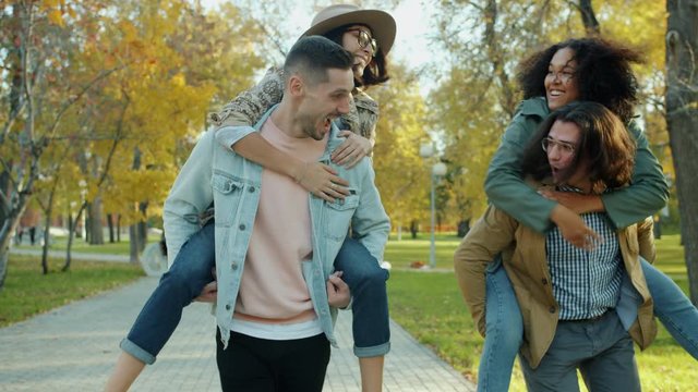 Joyful young men are giving piggyback ride to happy young women having fun doing high-five laughing in park. Friendship, youth and couples concept.
