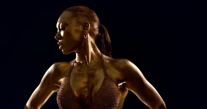 beautiful african sportswoman is posing in darkness, dressed in bra, skin covered by gold dye