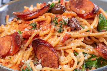 A bowl of chef prepared spaghetti with chorizo, black olives, and spinach in a creamy tomato sauce in a ceramic bowl on a wooden chopping board with red and white napery.