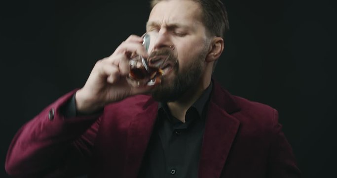 Brutal bearded man in expensive suit drinking whiskey and frowning, bad taste, poor quality