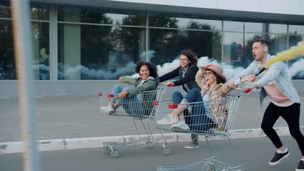 Slow motion of happy young people riding shopping carts holding smoke grenades laughing enjoying freedom in city. Lifestyle and emotions concept. - Powered by Adobe