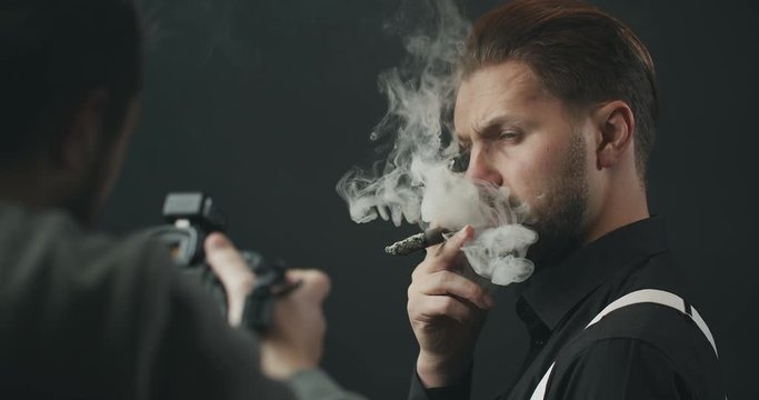 Photographer making shots of masculine bearded male in black shirt and white suspenders smoking cigar, fancy photo-session