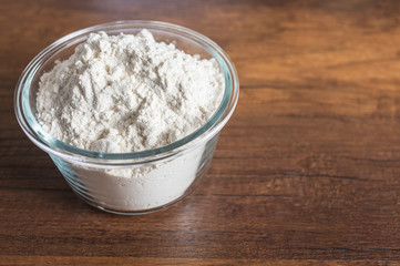 White flour in a glass bowl container on wood table surface.