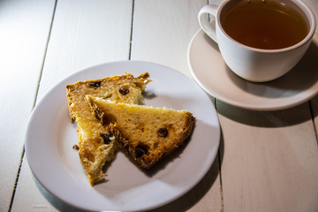 Two pieces of cake with raisins on a white plate and a white ceramic cup with tea on a white table from painted boards.