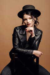 young beauty fashion model woman in  hat portrait on a beige background, black leather jacket,  sitting on a chair