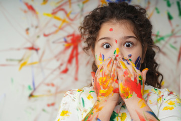 portrait of smiling little girl looking through her colorful hands and cheek  painted in kids room .  fear