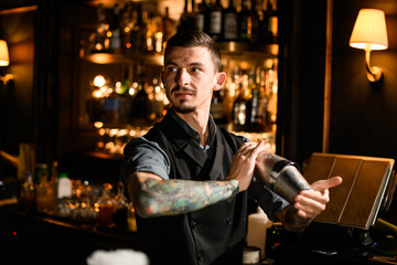 Tattooed bartender holding in hands a steel shaker with a deliciouds cocktail