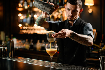 Male bartender pouring a smooth orange alcoholic cocktail drink from the steel shaker through the sieve