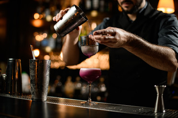 Male bartender pouring a crimson alcoholic cocktail drink from the steel shaker through the sieve