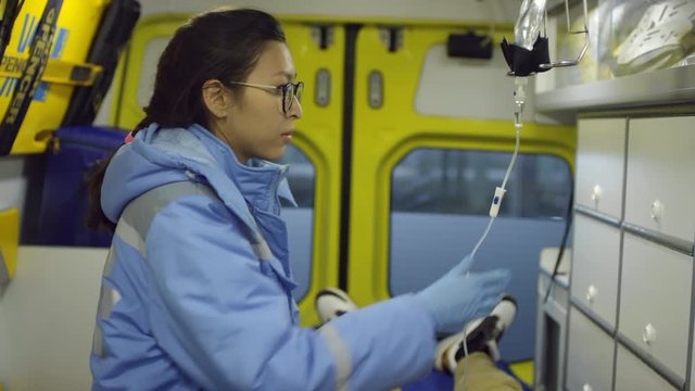 Handheld shot of busy female paramedic in glasses adjusting IV drop while working with patient in ambulance, then looking at camera
