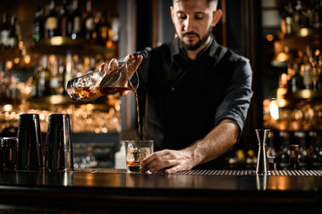 Bartender pouring a brown alcoholic drink from the measuring cup with straner to a glass