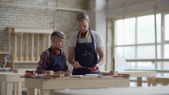Medium panning shot of middle aged carpenter explaining to son how to use chisel and stainless steel ruler standing at workbench in woodworking workshop