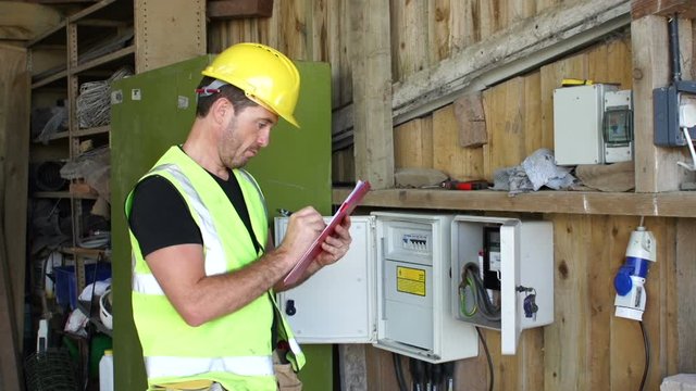Electrician inspects Fuse box and is using a Clipboard. The technician is wearing a hard hat and hi vis jacket - Stock Video clip footage. 