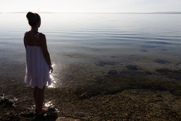 Back lit, silhouetted young woman in white dress with view of beautiful, pristine coastal bay on a glassy calm morning in South Australia.