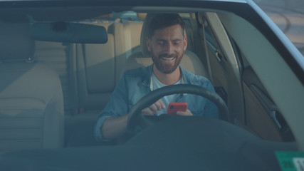 Attractive businessman texting in social network on smartphone sitting in modern car. Profile of handsome sociable guy with mobile phone planning a route through city.