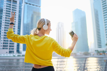 Sport and music. Happy young woman with earphones and smartphone dancing while exercising on city embankment.
