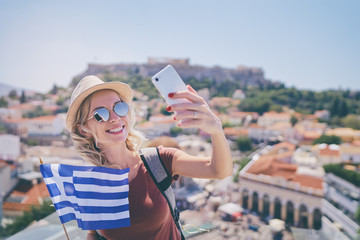 Enjoying vacation in Greece. Young traveling woman with national greek flag taking selfie on view of Athens city and Acropolis.