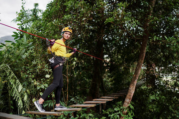 Naklejka na ściany i meble Young woman with climbing gear in an adventure extreme park climbing or passing on the rope road.