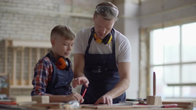 Rack focus of middle aged woodworker working in carpentry and showing little son how to make marks on wooden plank using pencil and triangle ruler