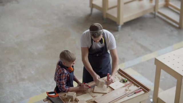 From above view of father and little son woodworking together at bench in carpentry shop. Dad teaching kid how to use chisel and wooden mallet