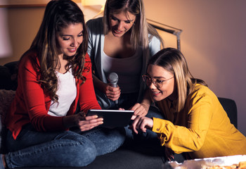 Three female friends having fun sitting on sofa playing karaoke at home.