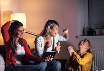 Three female friends having fun sitting on sofa playing karaoke at home.