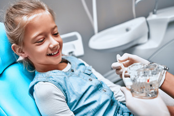 Pediatric dentist educating a smiling little girl about proper tooth-brushing, demonstrating on a model.