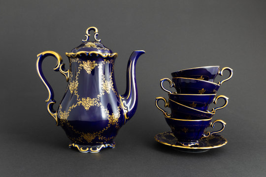 Closeup of a beautiful cobalt blue colored vintage porcelain tea set with golden floral pattern on dark gray background. The set includes a tea pot and a stack of tea cups.