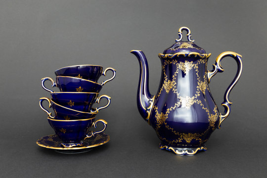 Closeup of a beautiful cobalt blue colored vintage porcelain tea set with golden floral pattern on dark gray background. The set includes a tea pot and a stack of tea cups.