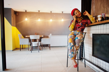 Enthusiastic african american woman in trendy coloured outfit with red beret chilling in cozy cafe, speaking on mobile phone.