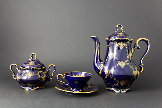 Closeup of a beautiful cobalt blue colored vintage porcelain tea set with golden floral pattern on dark gray background. The set includes a tea pot, a sugar bowl and a tea cup.