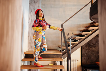 Enthusiastic african american woman in trendy coloured outfit with red beret chilling in cozy cafe, standing on stairs with cup of hot drink in hands.