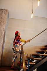 Enthusiastic african american woman in trendy coloured outfit with red beret chilling in cozy cafe, standing on stairs with cup of hot drink in hands.