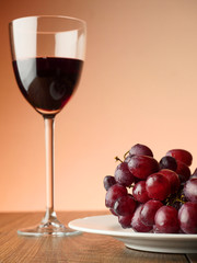 Glass of red wine and plate with red grapes on a wooden table, Warm light background. Grapes are in focus.