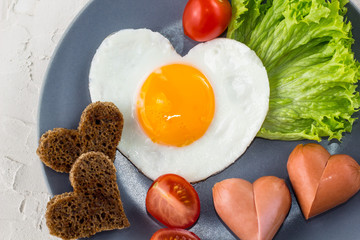 Fried eggs in the shape of a heart with sausages, herbs and tomatoes. Breakfast on Valentine's day on white background