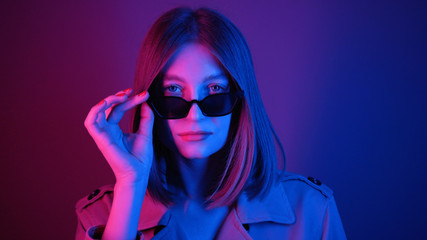 Classy young woman in sunglasses looking at camera, smiling. Shooting of adorable girl in stylish outfit. Good mood. Neon lights. Purple, colourful. Idea.