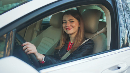 Fabulous Caucasian girl with brown hair sitting in driver's seat in new car, looking at camera and smiling. Portrait of amazing businesswoman. City life.