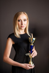 Beautiful young successful blonde woman with gold Cup winner on dark background