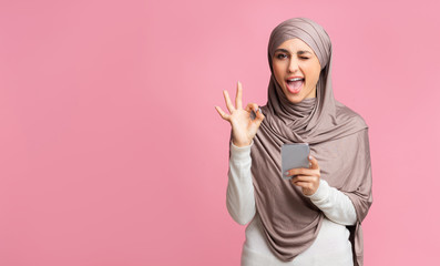 Arabic woman in hijab using smartphone, winking and showing ok gesture