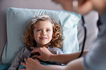 Unrecognizable female doctor examining small girl in bed in hospital.