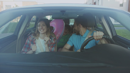 Front view of pretty married cuple and their kids riding a car in the city. Cheerful dad and his children giving high five having fun in the car during a family roadtrip to the seaside.