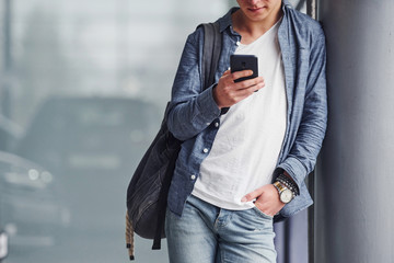 Hipster guy in nice clothes stands indoors against grey background with phone in hand
