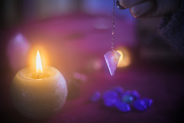 Young woman holding an amethyst pendulum with an esoteric background, candles, stones and crystals