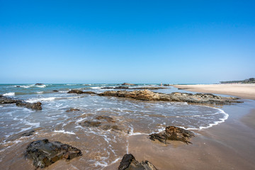 Fototapeta premium Long sandy beach background. Panoramic view of the Arabian sea coast in India. Large boulders on the shore. Surf Waves breaking on the stones. Horizon line of the ocean. 