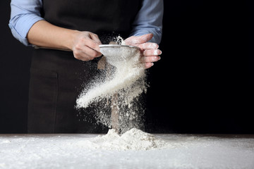 Woman sifting wheat flour at table against black background, closeup