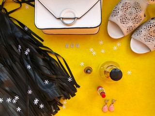 Party style flat lay. Female fashion. Black leather skirt with fringe, white clutch, white shoes on heels, wow text, perfume, red lipstick, red nail valish, earings, tinsels on yellow background. Copy