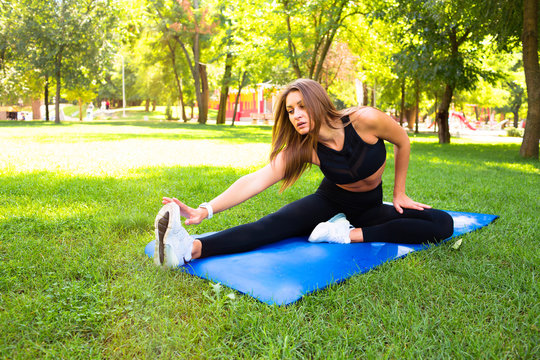 Image of smiling fitness woman 20s wearing headphones working out and stretching legs while sitting on exercise mat in green park