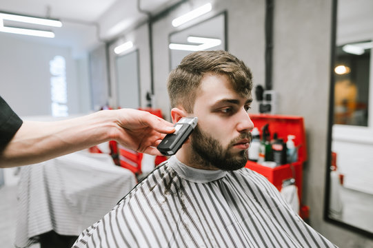 Hairdresser's hand cuts the man's beard trimmer. Portrait of bearded man sitting in chair in barbershop and doing hairstyle. Creating a male haircut at a men's hair salon.